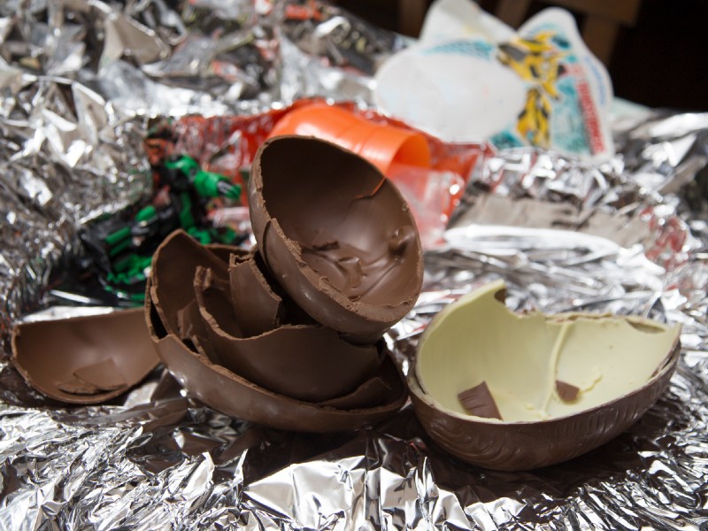Foto de ovo de chocolate. São várias cascas de ovo de Páscoa quebrados sobrepostas sobre um papel laminado. Uma das cascas é de chocolate branco e as demais são de chocolate preto